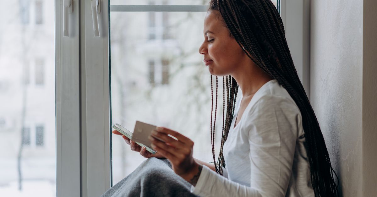 person sitting by a window at sunset with a phone showing a prayer app, looking contemplative