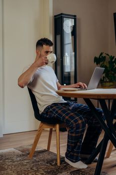 Man in pajamas drinks coffee while working on laptop at home office.