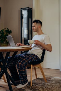 A man in casual home wear works on a laptop while enjoying a coffee. Modern lifestyle.