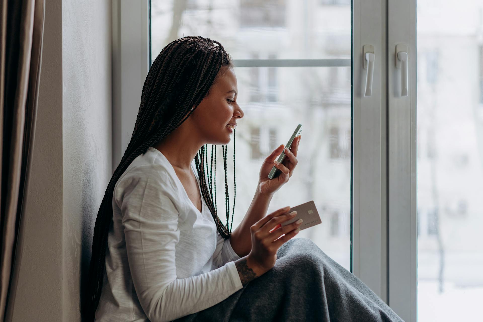 Woman sitting by the window shopping online with her phone and credit card, enjoying a relaxed day indoors.