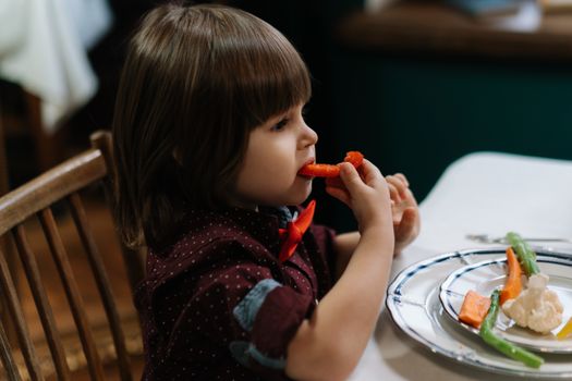 A toddler in a bow tie is eating vegetables such as carrot and cauliflower at a dinner table indoors.