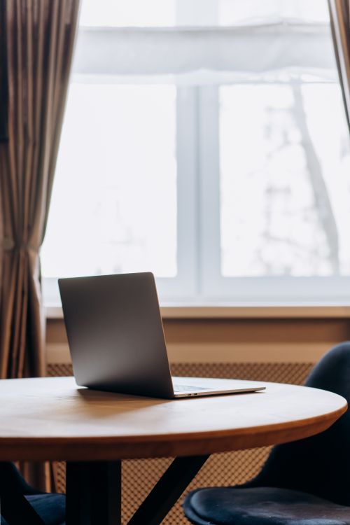 Free Image of a sleek laptop on a wooden round table near a window with curtains. Stock Photo