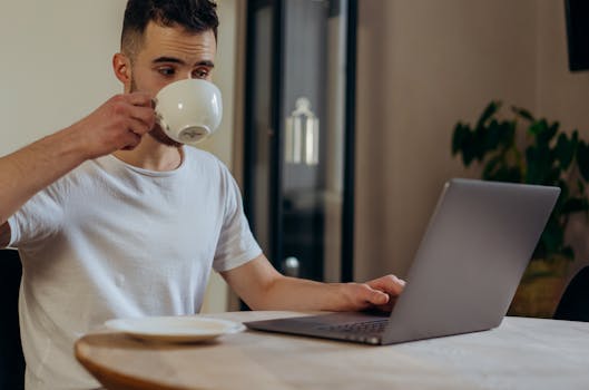 A man in a casual setting, sitting indoors, enjoying coffee while working on a laptop.