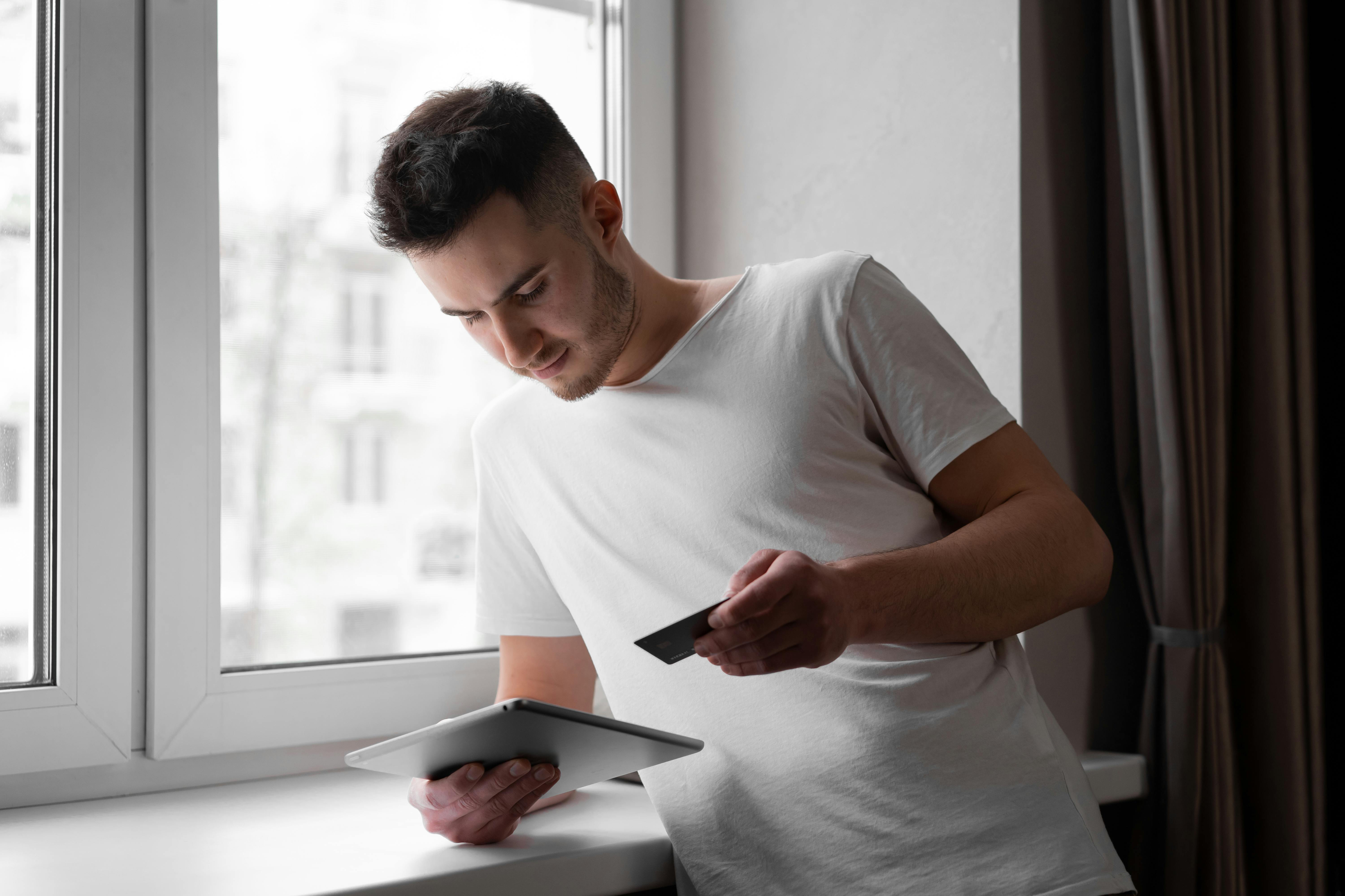 Man using digital tablet and credit card for online shopping by window indoors.