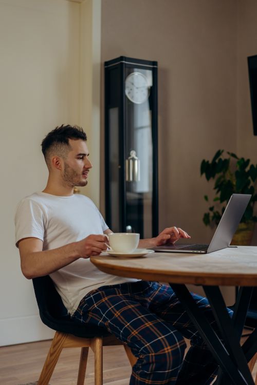 Free A man in checkered pants enjoys coffee while using a laptop at home. Calm indoor setting. Stock Photo