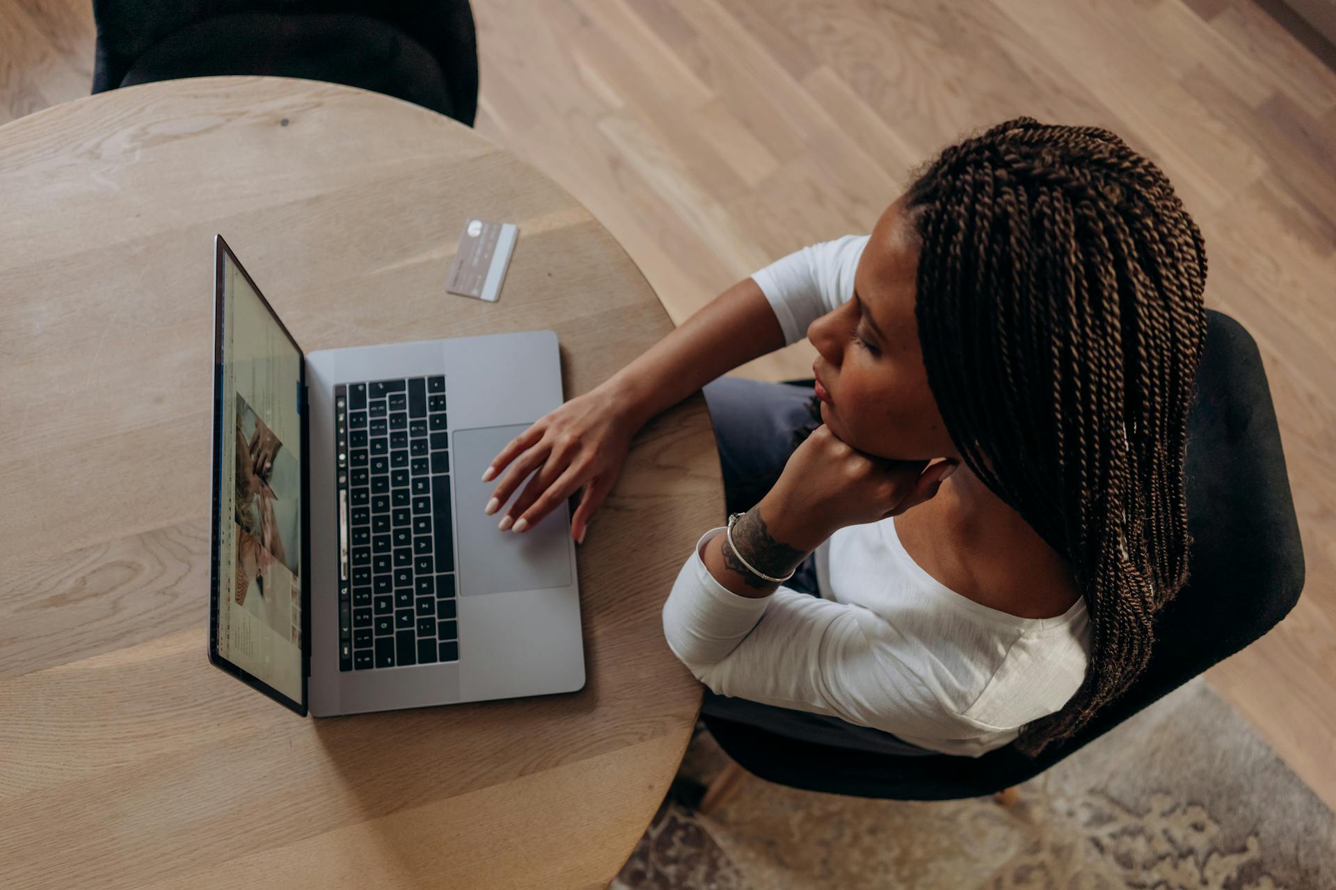 A woman sitting at a wooden table using a laptop with a credit card nearby, symbolizing online shopping or remote work.