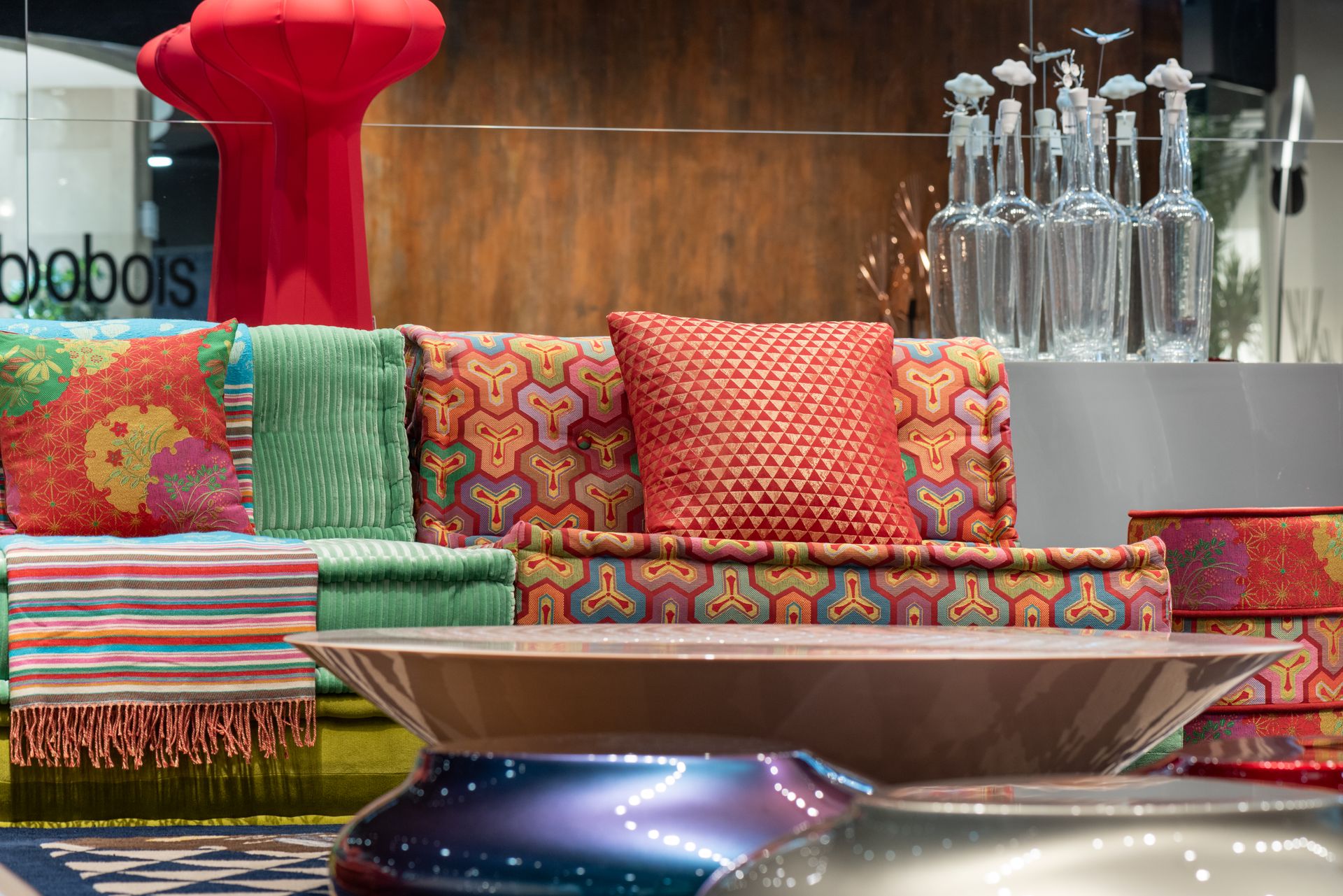 Multicolored comfortable sofa with bright cushions and vivid textile placed near modern round table in contemporary furniture store with glass wall and bottles