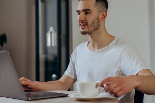A young man in a white shirt works on a laptop indoors, holding a coffee cup.