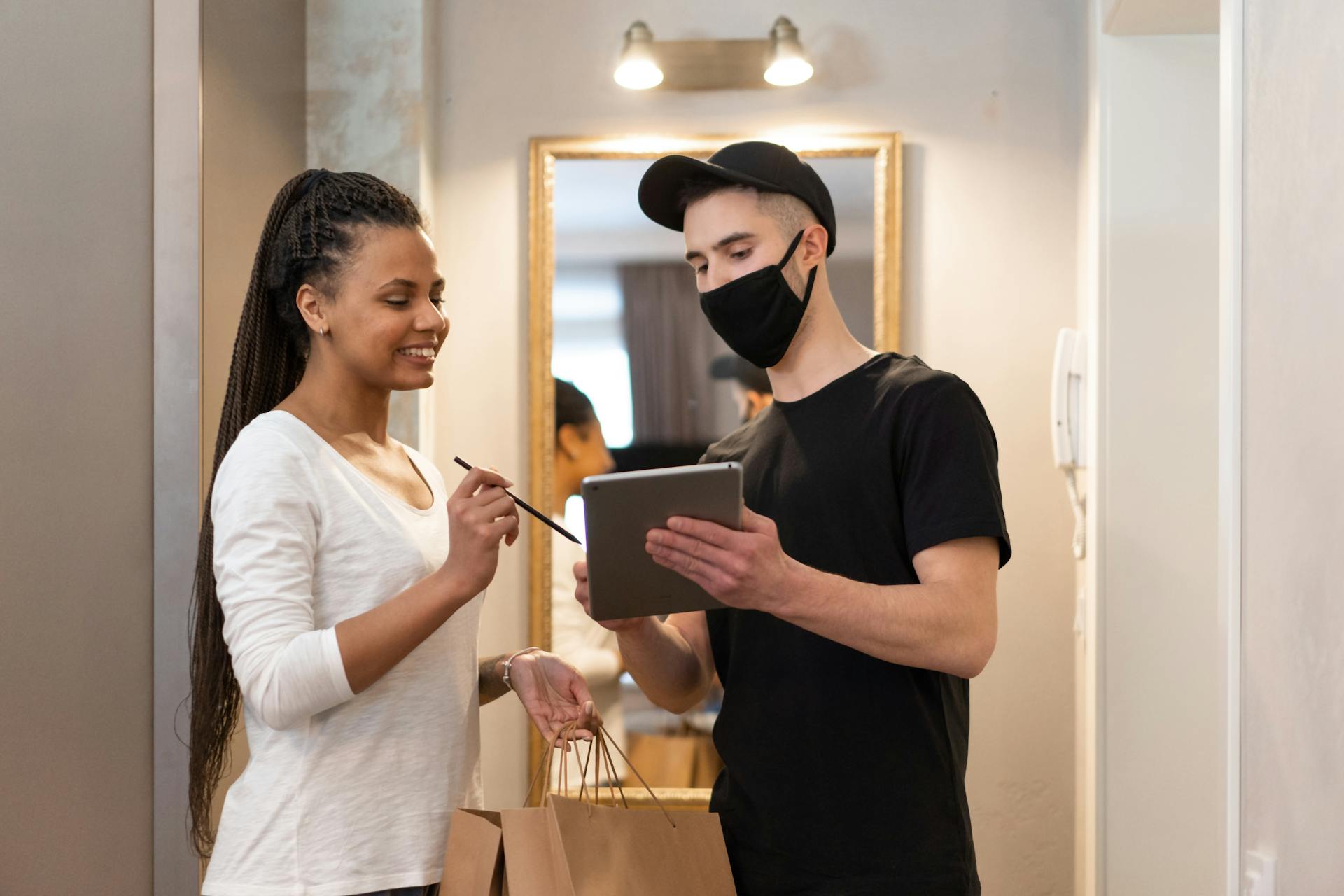 A delivery person and customer engage in a transaction at a home doorway, using a tablet for a seamless experience.