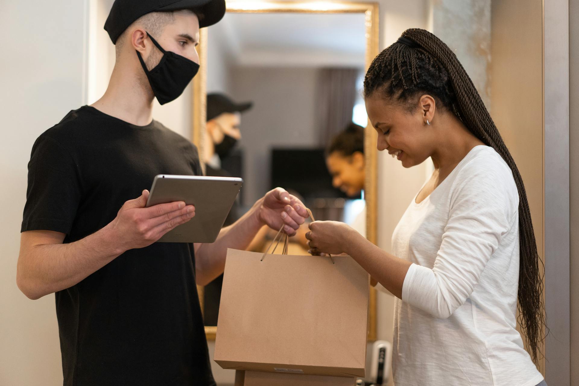 A deliveryman and a woman exchange a package indoors using a tablet for a cashless transaction.