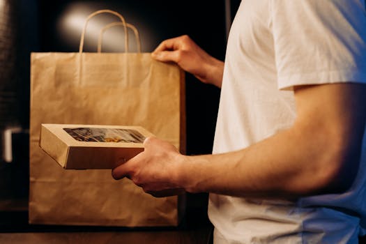 A person presenting a food delivery package and paper bag at home.