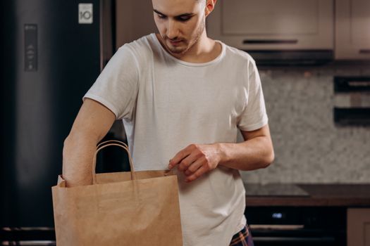 Male in white shirt unpacking groceries from paper bag in contemporary kitchen setting.