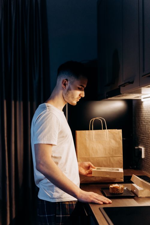 Free A young man examines a takeout box in a dimly lit modern kitchen, emphasizing a casual evening mood. Stock Photo