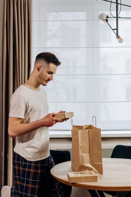 Free A man stands indoors opening a food delivery from a paper bag on a table. Stock Photo