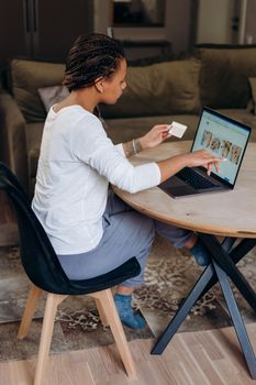 A woman uses a credit card for online shopping, sitting at a home dining table with a laptop.