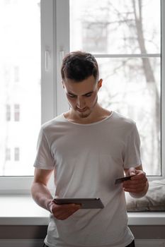 A young man uses a tablet and credit card for online shopping at home by a window.