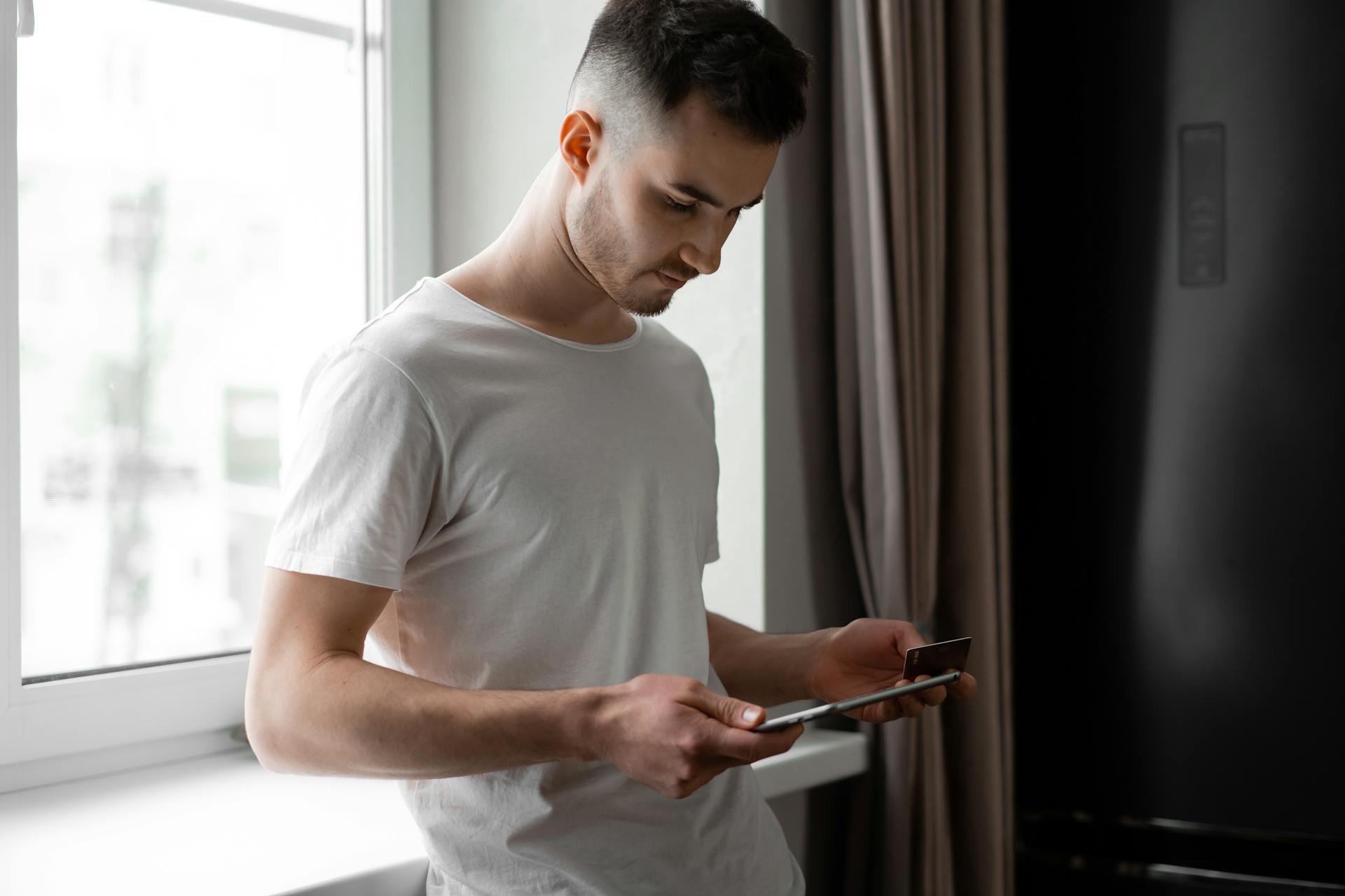 Young man standing by a window, using tablet and credit card for online shopping indoors.