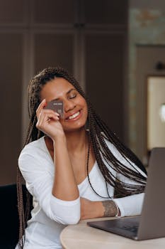Smiling woman indoors with long braids holding credit card near laptop, enjoying online shopping.
