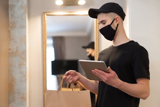 A deliveryman in a mask uses a tablet for contactless service indoors, holding paper bags.