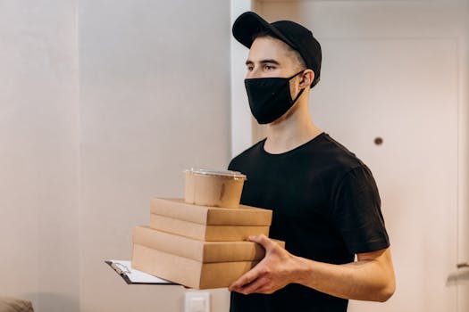 A delivery man in black attire with a mask holding food packages inside.