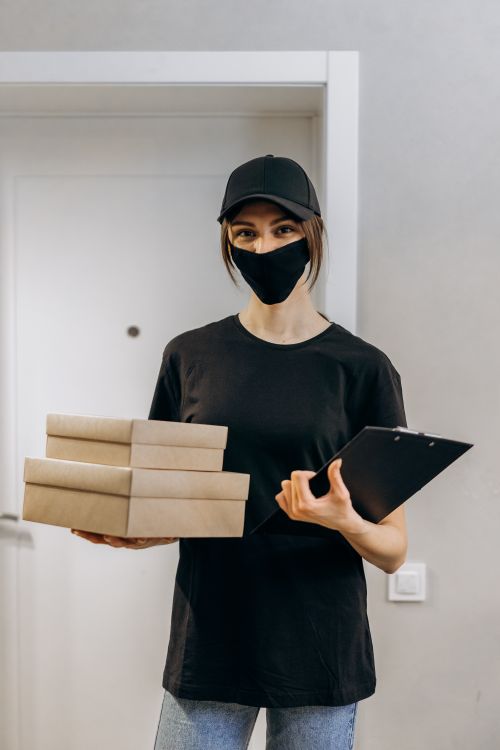 Free A delivery woman in a mask holds packages and a clipboard indoors, emphasizing safety. Stock Photo