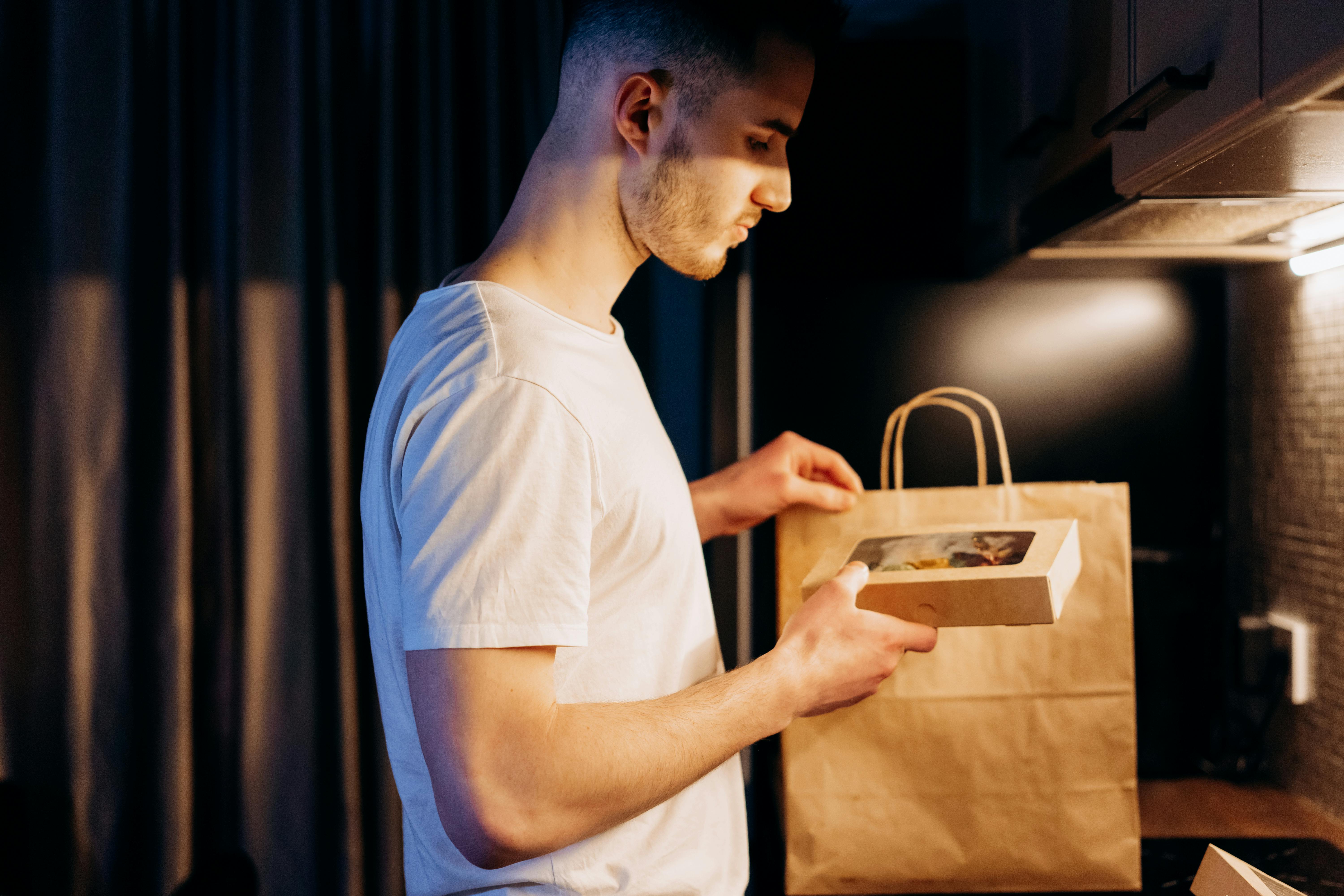 A man unpacks a food delivery from a paper bag in a warmly lit kitchen setting.
