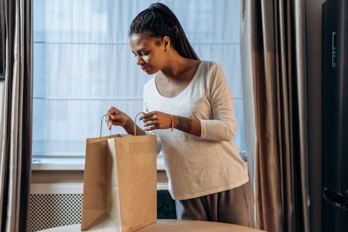 Free A woman looks inside a paper delivery bag at home, showing curiosity and anticipation. Stock Photo