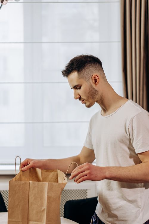 Free A young man in a white t-shirt examining a paper bag indoors. Modern setting with natural light. Stock Photo