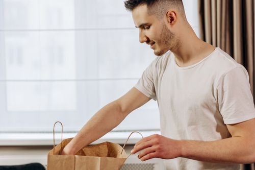 Free Young man in a white t-shirt smiles while unpacking a paper bag indoors, near a window. Stock Photo