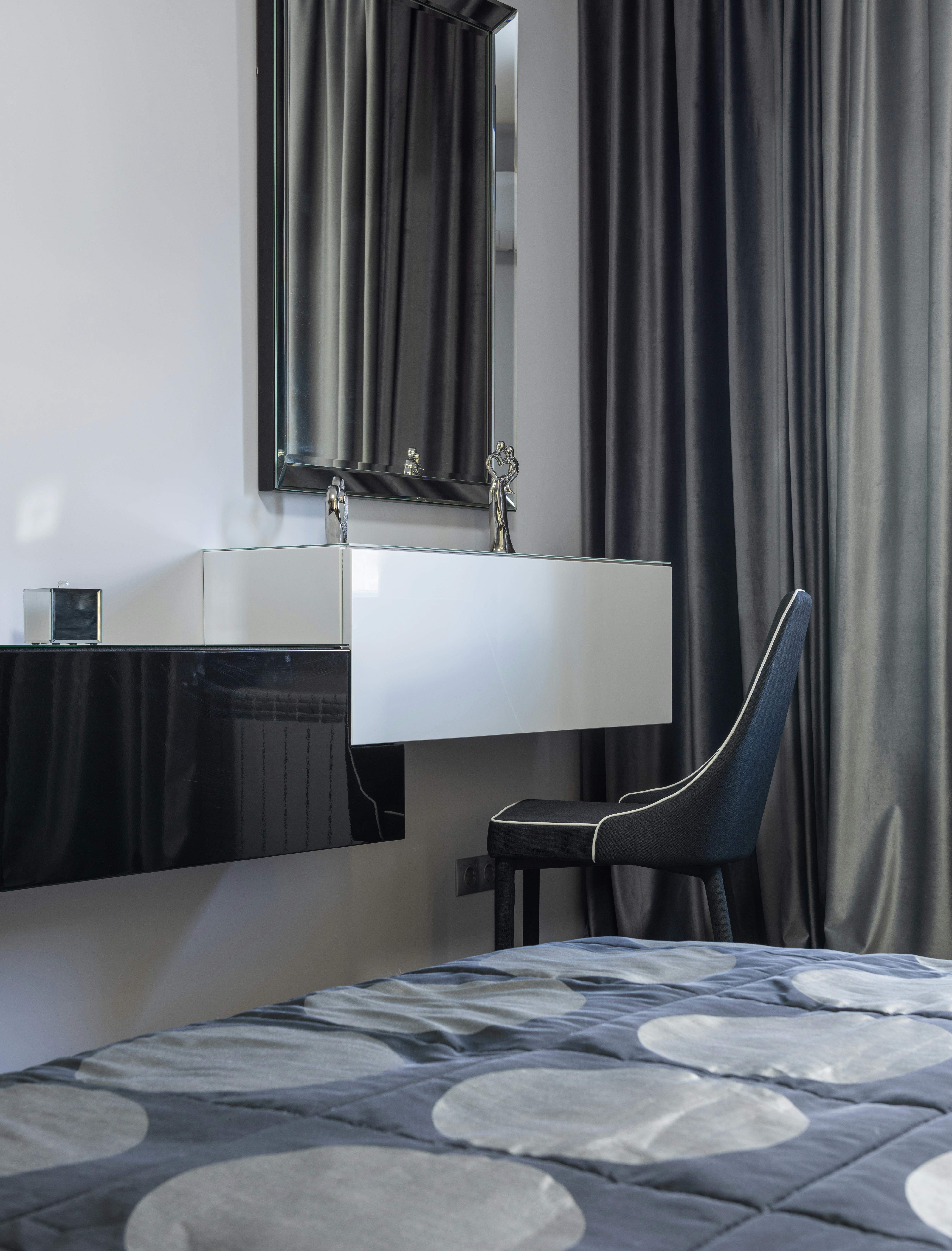 Free Bed with cover against chair and table with decorative statuettes under mirror reflecting curtain in house Stock Photo