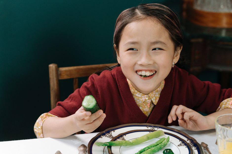 A happy child in a maroon sweater enjoying a meal with fresh vegetables at a dining table.