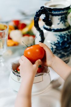 A child's hands holding a ripe tomato next to a vintage porcelain teapot, suggesting a fresh and healthy lifestyle.