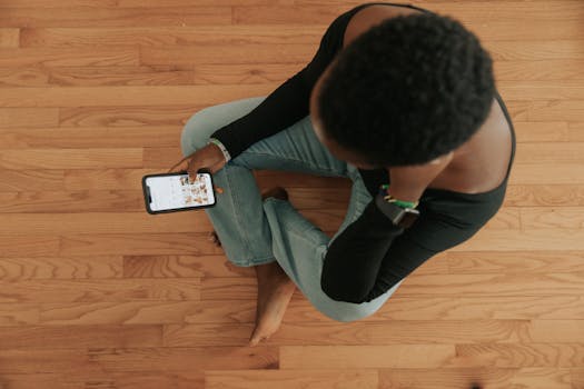 Person sits cross-legged on wooden floor, browsing on smartphone with focus on digital interaction.