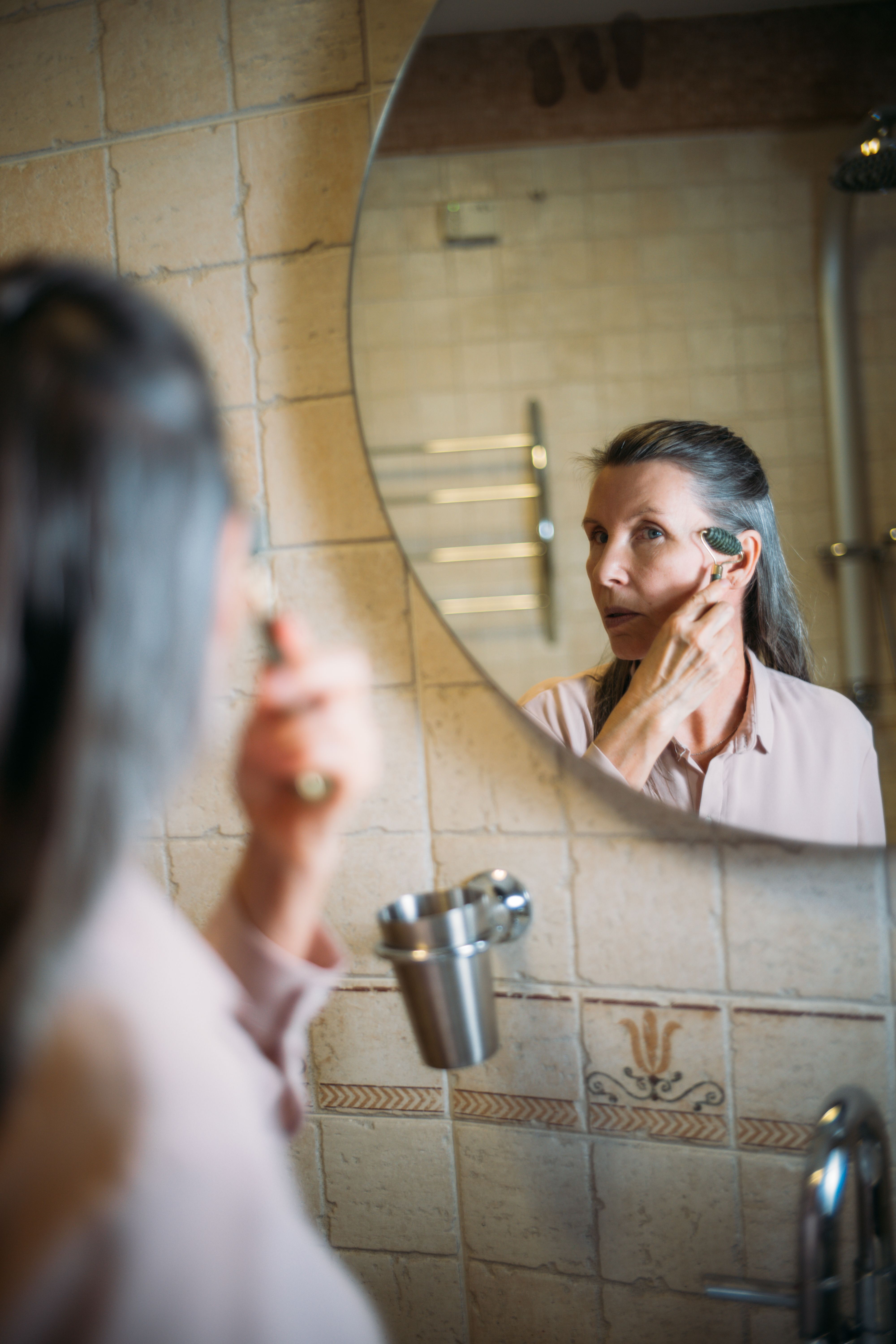 Elderly woman using a jade roller for facial care in the mirror, embracing self-care routine.