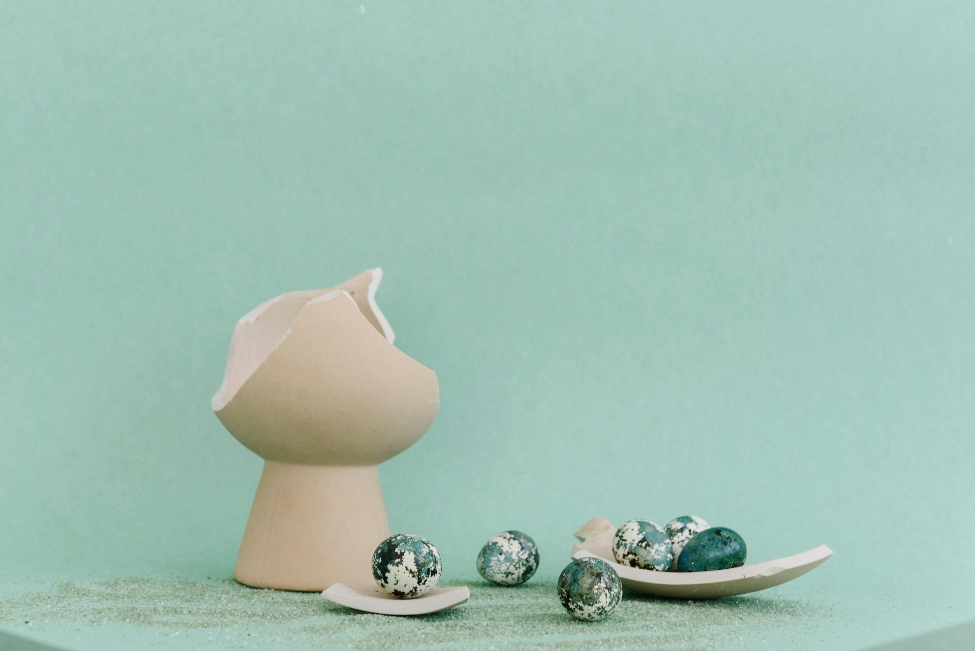 Artistic arrangement of a broken ceramic vase and speckled eggs on green.