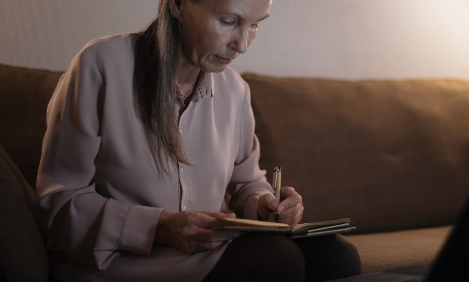A senior woman writing in a notebook while sitting on a couch indoors.