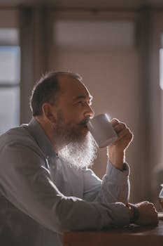Senior man with a beard enjoying a warm beverage indoors, bathed in soft daylight.