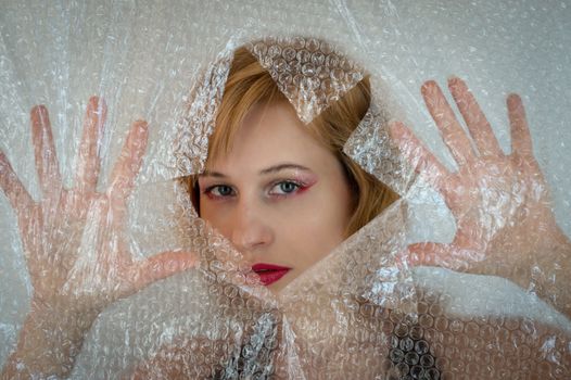 Attractive female model with bright makeup looking at camera through hole in transparent polythene package while standing in studio with raised hands
