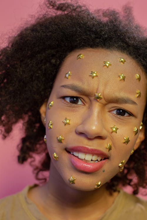 Resentful African American female teenager with yellow stars sequins on face looking at camera on pink background in modern studio