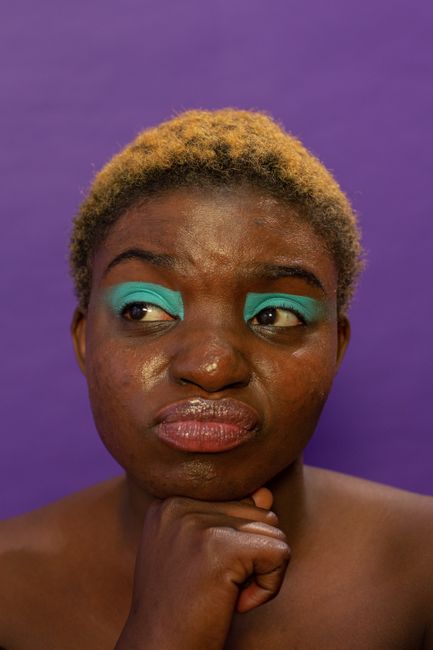 Doubting African American female model with dyed yellow hair and bright eyeshadow looking away on purple background in modern studio