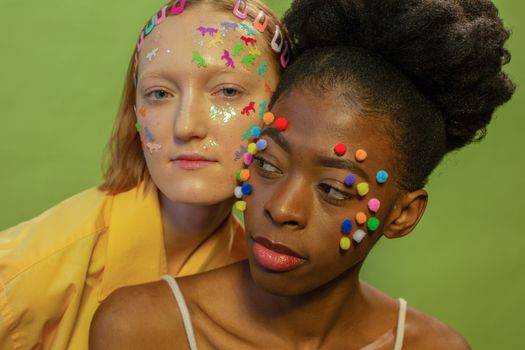 Stylish young multiethnic friends with bright stickers and pom pom balls on face on green background