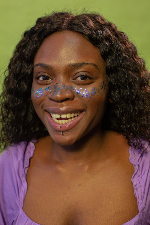 Cheerful African American female with sparkling glitter on face looking at camera on green background