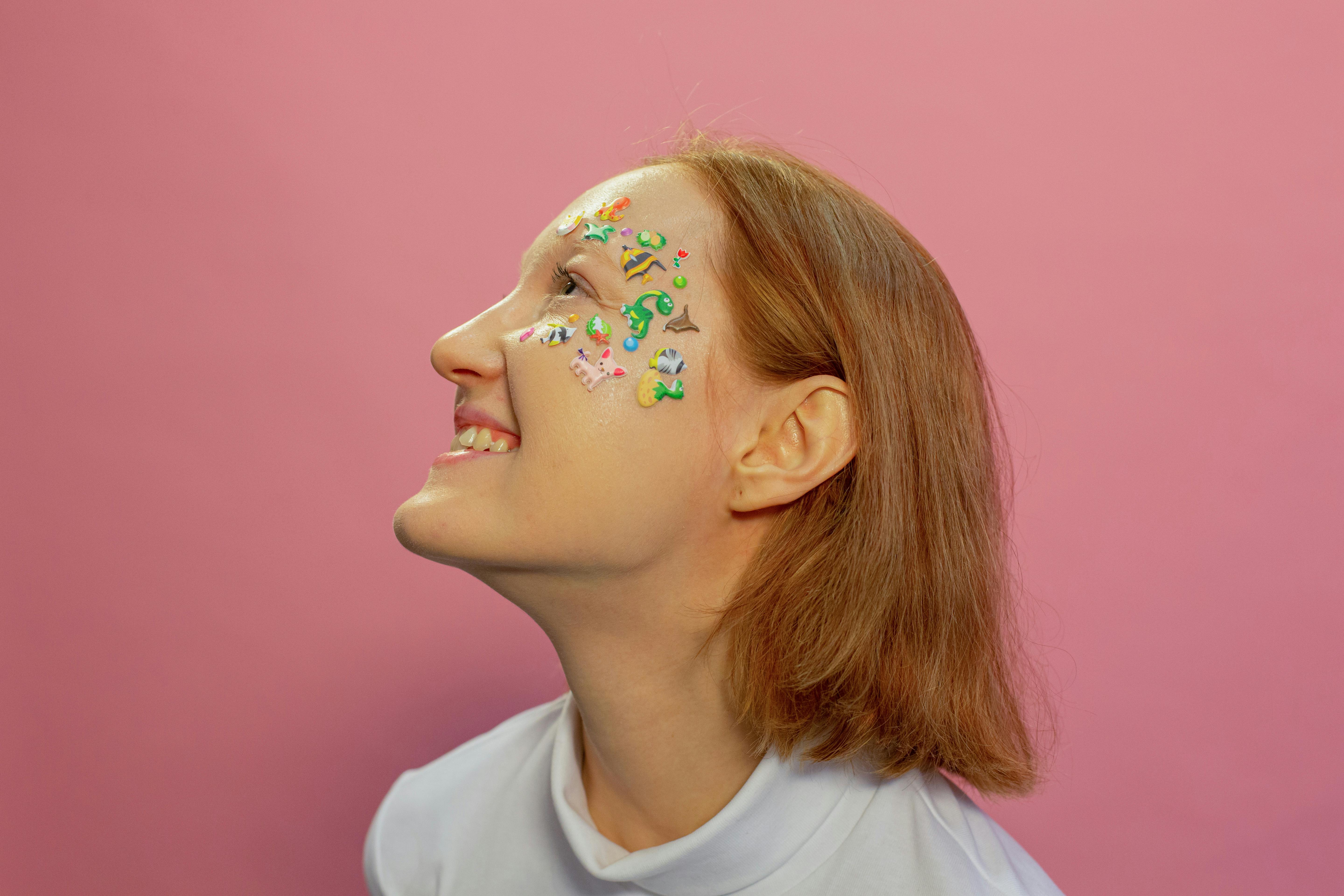 A captivating portrait of a woman smiling with colorful face stickers against a vibrant pink background.