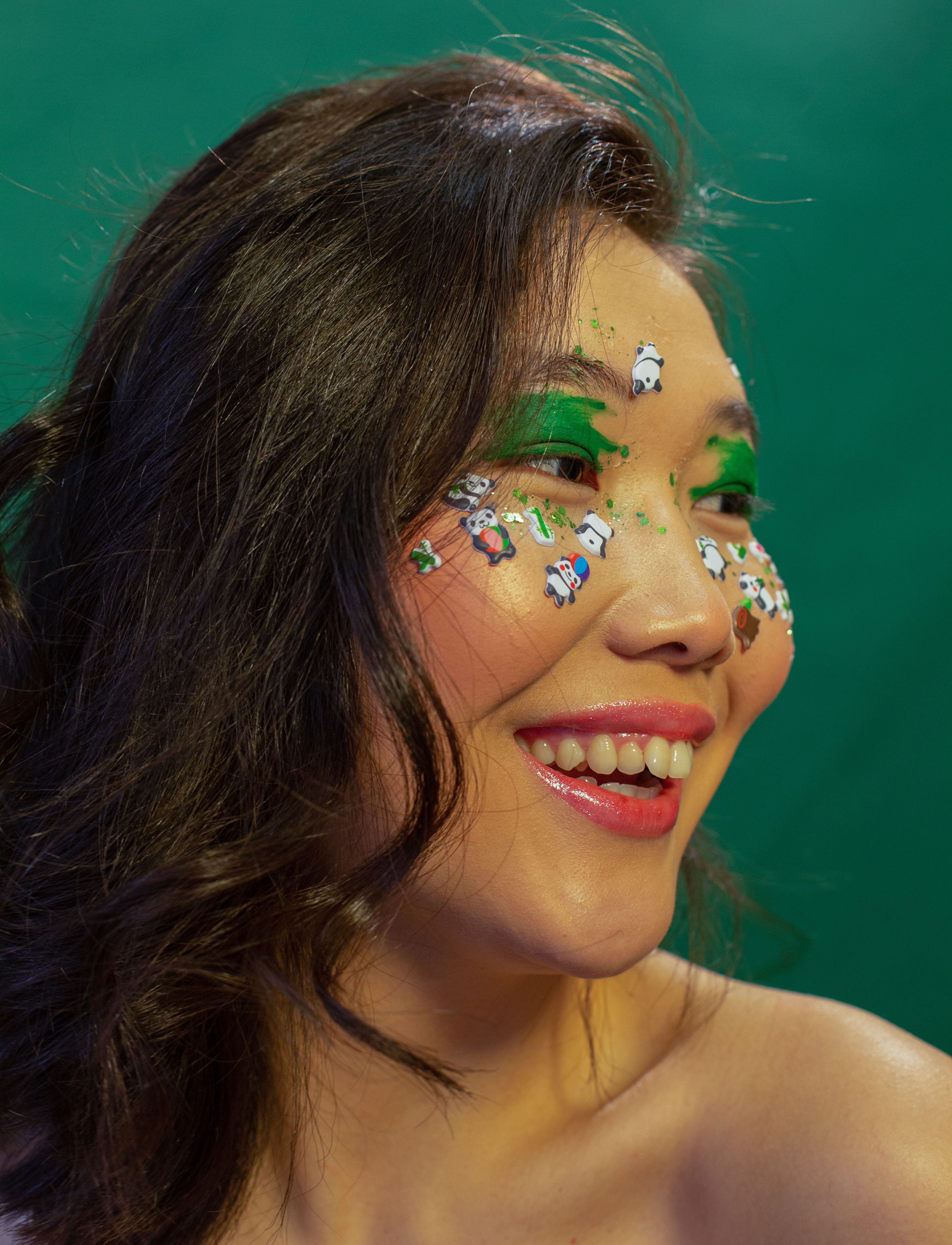 Asian woman smiling with green eye makeup and playful face stickers against a green background.