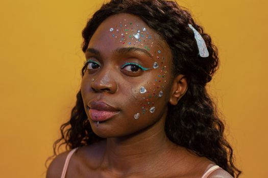 Close-up portrait of a young woman with artistic makeup and glitter on a vibrant yellow background.