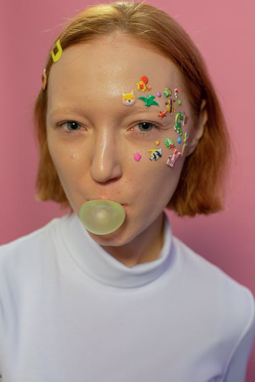 Young lady in casual clothes and colorful animal stickers on face and hairpins in hair looking at camera while blowing bubble with gum on pink background in light studio