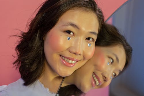 Smiling young ethnic lady in makeup looking at camera with shining rhinestones in shape of hearts on cheeks near mirror with self reflection on pink background in light studio