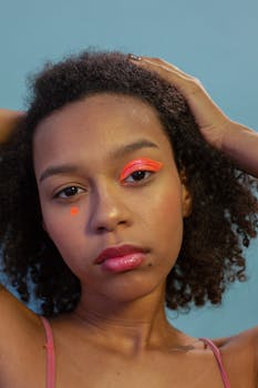 Serious young black teen with orange neon eyeshadow and dot on face looking at camera with hand on head in bight studio on blue background