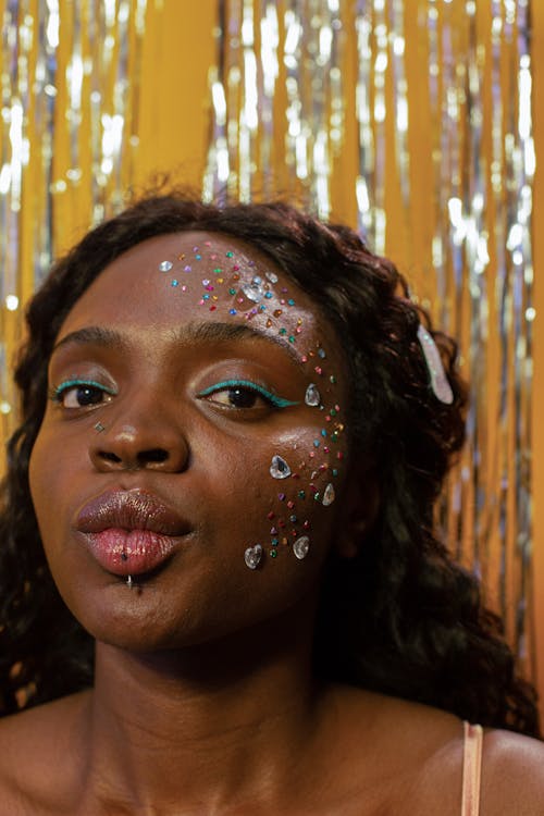 Free Young African American lady with makeup and piercing on lip with hearts rhinestones on face looking at camera while blowing air kiss near yellow wall with tinsel streamer Stock Photo