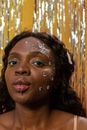 Young African American lady with makeup and piercing on lip with hearts rhinestones on face looking at camera while blowing air kiss near yellow wall with tinsel streamer